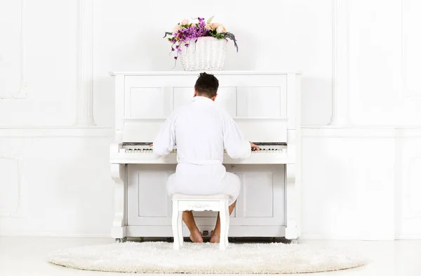 Man in bathrobe enjoys morning while playing piano. Talented musician concept. Man sleepy in bathrobe sit in front of piano musical instrument in white interior on background, rear view. — Stock Photo, Image