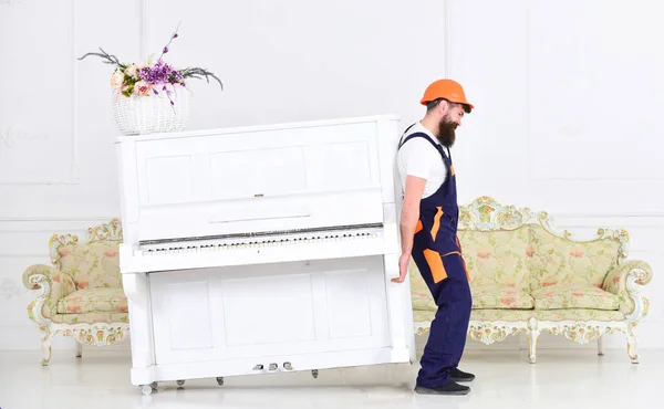 De lader beweegt piano-instrument. Koerier levert meubels, verhuist, verhuist. Zware lasten concept. Man met baard werknemer in helm en overall tilt, inspanningen om piano te bewegen, witte achtergrond — Stockfoto