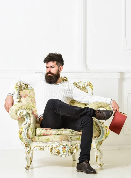 Hombre con barba y bigote se sienta en el sillón, sostiene libro, fondo blanco de la pared. Conocedor de cara reflexiva terminado libro de lectura. Reflexiones sobre el concepto de literatura. Chico pensando en literatura . —  Fotos de Stock