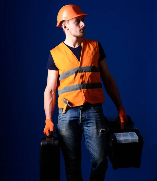 Mężczyzna w kasku, twardy kapelusz trzyma skrzynkę z narzędziami i walizkę z niebieskim tłem. Koncepcja profesjonalnego mechanika. Robotnik, złota rączka, mechanik, budowniczy na spokojnej twarzy niesie torby z profesjonalnymi narzędziami. — Zdjęcie stockowe