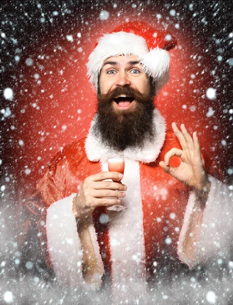 Stilig skäggig jultomte man med långt skägg på leende ansikte innehav glas alkoholhaltiga skott i jul eller jul tröja och nyår hatt och visar ok på röd studio backgrou — Stockfoto