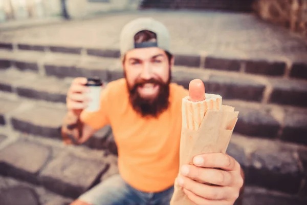 Despreocupado hipster comer junk food while sit on stairs. Um lanche de homem faminto. Comida de plástico. Um tipo a comer cachorros quentes. Homem barbudo mordida saborosa salsicha e beber copo de papel. Comida de rua tão boa. Estilo de vida urbano nutrição — Fotografia de Stock