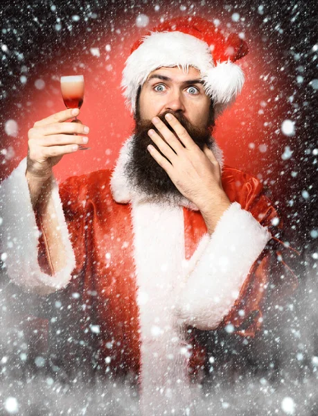 Stilig skäggig jultomte man med långt skägg på förvånad ansikte innehav glas alkohol skott i jul eller jul tröja och nyår hatt på röd studio backgrou — Stockfoto