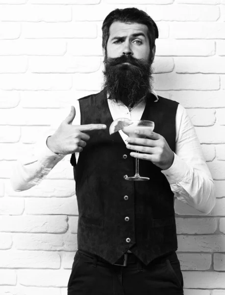 Handsome bearded man with long beard and mustache has stylish hair on serious face holding glass of alcoholic beverage in vintage suede leather waistcoat on white brick wall studio backgrou — Stock Photo, Image