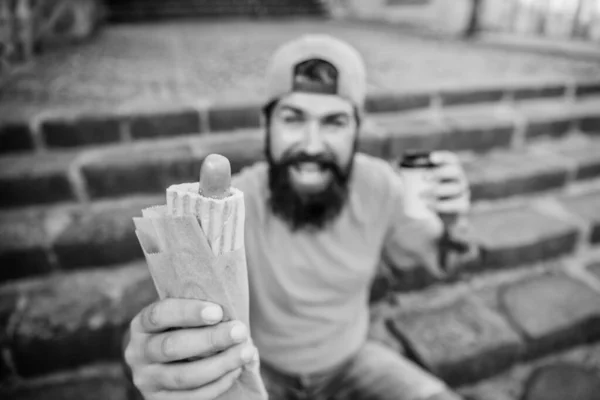 Despreocupado hipster comer comida chatarra mientras se sienta en las escaleras. Snack de hombre hambriento. Comida chatarra. Un tipo comiendo perritos calientes. Hombre barbudo bocado sabrosa salchicha y beber taza de papel. Comida callejera tan buena. Vida urbana nutrición —  Fotos de Stock