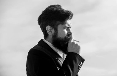 Sigaralı adam nikotin etkisinden hoşlanır. Sigara zevki ve nikotin bağımlılığı hakkındaki gerçekler. Sakallı ve bıyıklı bir adam sigara tutuyor. Sakallı hippi sigara içiyor mavi gökyüzü arka planı