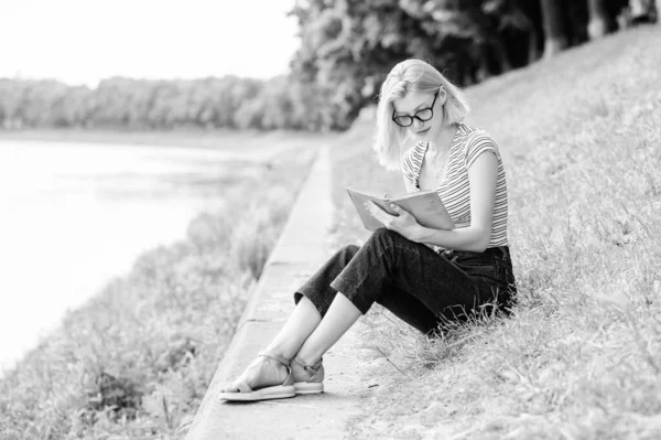 梦想度假的女人 暑假的文学。 休息和业余爱好。 暑假。 女孩坐在河边的绿草上看书。 3.女孩下班后在河边休息 — 图库照片
