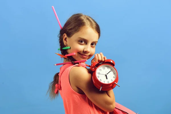 Niño en vestido rosa con lápices de colores sostiene reloj despertador — Foto de Stock