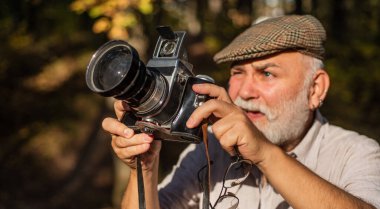Profesyonel Doğa Fotoğrafçısı. Sakallı emekliler seyahat fotoğrafları çeker. Eski model fotoğraf makinesi. Sakallı, olgun bir erkek. Retro film kamerasıyla fotoğraf çekmek. Eski model fotoğraf makinesi açık havada.