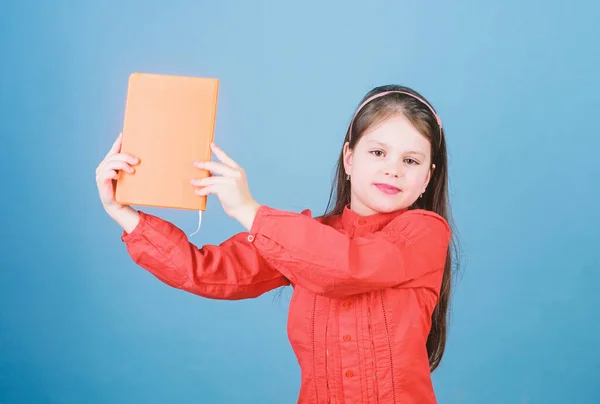 Mira este libro. Un pequeño alumno tomó el libro de la biblioteca. Niño adorable de la escuela con libro de actividades. Linda niña sosteniendo el cuaderno con cubierta naranja, espacio para copiar — Foto de Stock