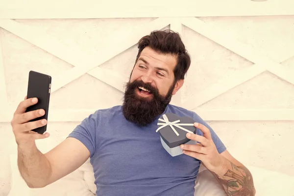 Good morning. Special gift. Bearded man using mobile technology in bed. Handsome guy taking selfie with gift box. Happy selfie. Modern life new technology. Technology concept. Pleasant conversation
