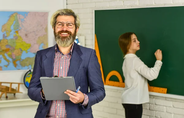 Szczęśliwy nauczyciel i dziecko w klasie. zmiana obowiązków. Brodaty człowiek robi notatki w schowku. Mała dziewczynka pisze odpowiedź na tablicy. egzamin lub lekcja. Z powrotem do szkoły. nowoczesna koncepcja edukacji — Zdjęcie stockowe