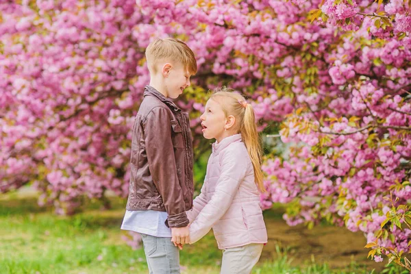 Love is in the air. Tender love feelings. Little girl and boy. Romantic date in park. Spring time to fall in love. Kids in love pink cherry blossom. Couple adorable lovely kids walk sakura garden — Stock Photo, Image