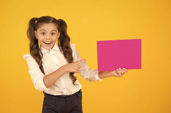Apresentando seu produto. Menina feliz apontando o dedo para papel rosa em branco para publicidade de produtos em fundo amarelo. De volta ao produto escolar. Chamando a atenção para o produto ou evento, espaço de cópia — Fotografia de Stock