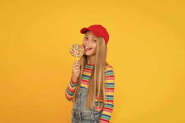 Lutscher macht sie glücklich. niedliches kleines Mädchen, das Regenbogen-Süßigkeiten auf gelbem Hintergrund isst. kleines Kind leckt Bonbons am Stock. entzückendes glückliches Kind beim Dessert mit Zuckerbonbons. Süßwarenladen — Stockfoto