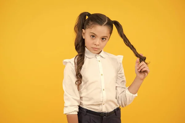 Кудрявые хвостики для возвращения в школу. Очаровательная маленькая девочка скручивает волосы вокруг пальца на желтом фоне. Милый маленький ребенок с длинными брюнетками. Стиль волос для школы — стоковое фото