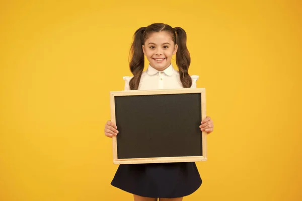 Um pequeno publicitário. Uma criança a segurar um quadro negro no fundo amarelo. Criança com quadro preto para propaganda escolar. Menina com quadro vazio para o seu texto, espaço de cópia — Fotografia de Stock