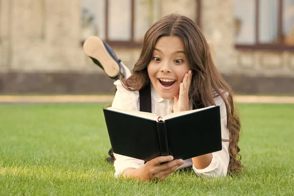 Boek met onverwachte verrassing. Verrast kind lees boek over groen gras. Meisje leest met verrassende emotie op het gezicht. Schattig klein schoolmeisje houden mond open van grote verrassing. Verrassingsconcept — Stockfoto