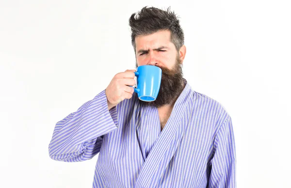 Мачо в халате пьет чай или кофе со спящим лицом. Парень выглядит сонным, ему нужен кофе. Человек с бородой и усами держит кружку кофе, изолированного на белом. Утренний кофе . — стоковое фото
