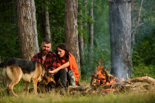 Paar spielt mit Schäferhund am Lagerfeuer, Waldhintergrund. — Stockfoto