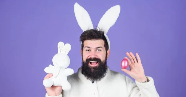 Diversão de Páscoa. Homem barbudo em traje de coelho com ovo de Páscoa e brinquedo de lebre. Primavera, nova vida e fertilidade. Homem feliz com orelhas de coelho segurando coelho brinquedo e ovo. Celebração de férias de primavera — Fotografia de Stock