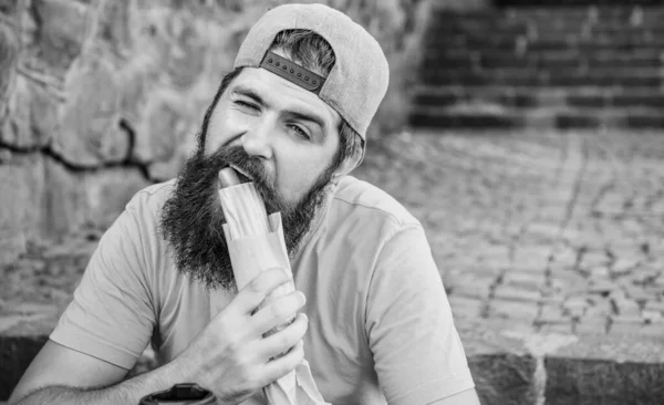 Hipster insouciant manger de la malbouffe tout en s'asseyant sur les escaliers. Un casse-croûte affamé. De la malbouffe. Un type qui mange du hot dog. Homme barbu mordre savoureuse saucisse et boire tasse en papier. La nourriture de rue est si bonne. Mode de vie urbain nutrition — Photo