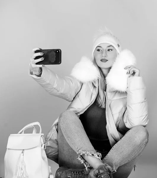 girl in puffed coat make selfie. faux fur fashion. warm winter clothing. phone selfie. flu and cold season. Leather bag fashion. woman in beanie hat with backpack. happy winter holidays. web blogger