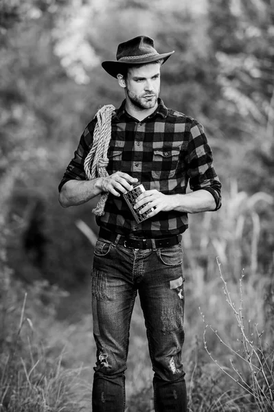 Whisky Bourbon. Cultura occidental. El hombre con sombrero sostiene la cuerda y el frasco. Herramienta Lasso vaquero americano. Brutal vaquero bebiendo alcohol. Hombre hermoso vaquero naturaleza fondo. Trabajador de rancho vaquero —  Fotos de Stock