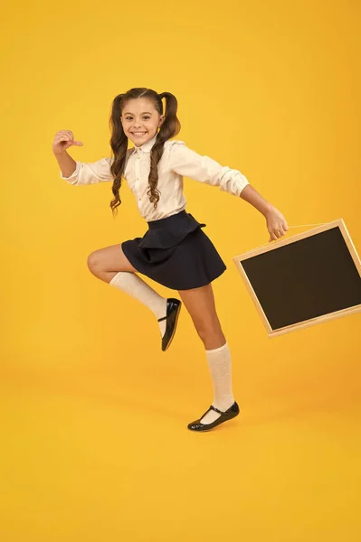 Começar a escola com um sorriso. Criança pequena feliz indo para a escola em fundo amarelo. Menina ativa em uniforme escolar segurando quadro-negro. Voltar à época escolar, espaço de cópia — Fotografia de Stock