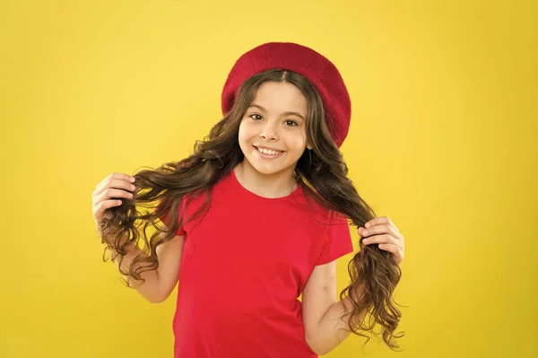 Glücklich und schön. Kind glücklich niedliches Gesicht liebenswert lockiges Haar gelb Hintergrund. Beauty-Tipps für gepflegtes Haar. Lächelndes Kind. Kleine Mädchen mit langen gesunden, glänzenden Haaren tragen einen roten Hut. Kleines Mädchen mit langen Haaren — Stockfoto