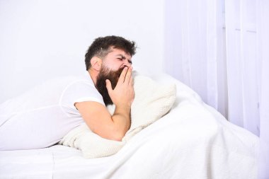 Gömlekli bir adam yatakta uzanırken esniyor, arka planda beyaz bir duvar ve perde var. Uykucu konsepti. Uykulu yorgun suratlı adam esniyor. Sakallı ve bıyıklı maço. Esniyor, dinleniyor, dinleniyor..