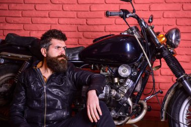 Hipster, düşünceli, deri ceketli vahşi motorcu oturur, motosiklete yaslanır. Sakallı bir adam, deri ceketli bir motorsikletli, garajda, tuğla duvarlı arka planda. Bisikletçilerin yaşam tarzı konsepti.