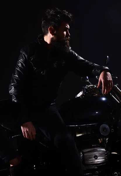 Homme à barbe, motard en veste de cuir s'appuie sur la moto dans l'obscurité, fond noir. Macho, motard brutal en veste en cuir se tient près de la moto la nuit. Brutalité et concept masculin . — Photo