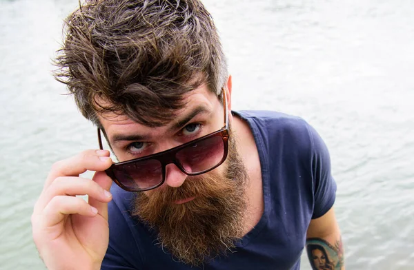 Homme avec barbe et moustache regardant hors des lunettes de soleil, rivière sur fond. Hipster sur le visage confiant avec des lunettes de soleil à la mode. Concept de mode masculine. Guy semble confiant avec des lunettes de soleil élégantes — Photo