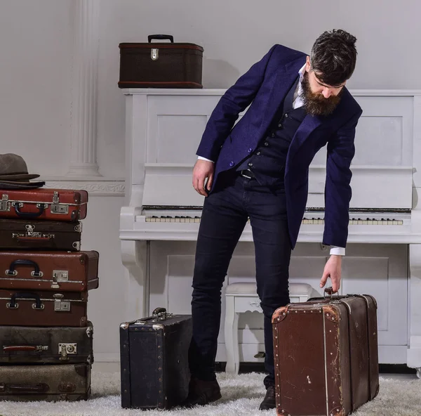 Concepto de entrega de equipaje. Hombre, viajero con barba y bigote con equipaje, lujoso fondo interior blanco. Macho elegante en la cara estricta se encuentra cerca de la pila de maleta vintage, sostiene la maleta . — Foto de Stock