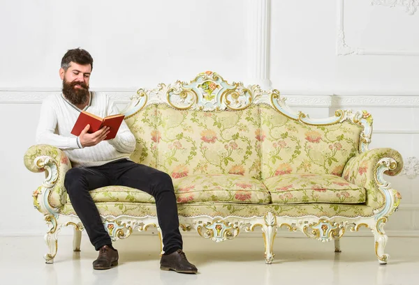 Concepto de literatura humorística. Un tipo leyendo un libro viejo con placer. Macho en el sonriente libro de lectura de caras. Hombre con barba y bigote se sienta en el sofá de estilo barroco, sostiene libro, fondo blanco de la pared . —  Fotos de Stock