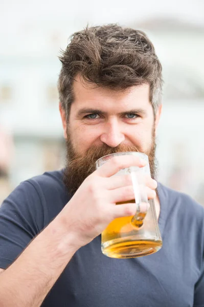 Skäggig man håller ölmugg, dricker öl utomhus. Koncept för hantverksöl. Mannen med långt skägg ser avslappnad ut. Man med skägg och mustasch på glad leende ansikte, ljus bakgrund, defocused. — Stockfoto