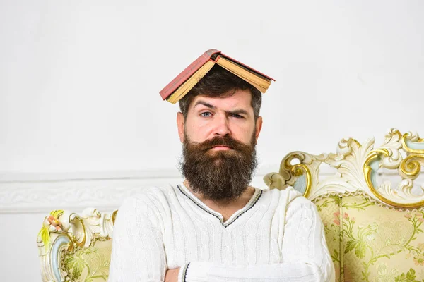Macho nyitott könyv a fején, mint a tető. Túlhajszolja magát. Guy, a tanár túl sokat tanított, és őrült professzor lett. Férfi szakállal és bajusszal ül a kanapén, fehér fal háttér. — Stock Fotó