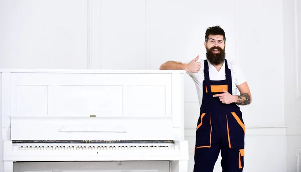 Man with beard and mustache, worker in overalls lean on piano, white background. Loader shows thumb up gesture. Courier delivers furniture in case of move out, relocation. Delivery service concept.