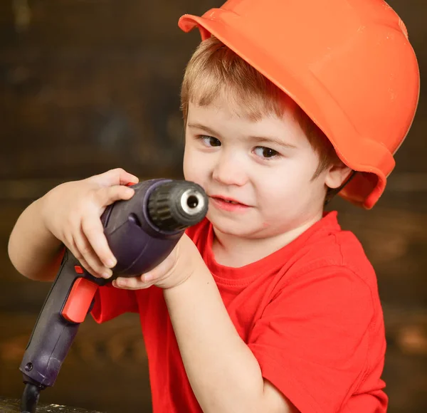Rapaz loiro de close-up com broca grande. Um pequeno construtor no estaleiro. Criança bonito em capacete protetor laranja isolado no fundo marrom desfocado — Fotografia de Stock