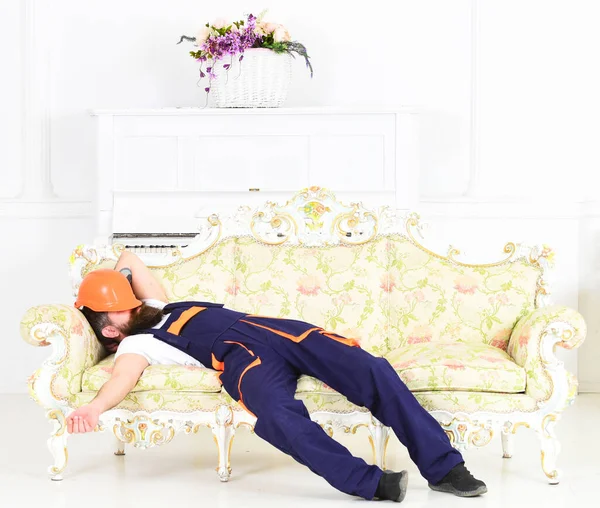 Trabajador barbudo durmiendo en sofá en habitación blanca con casco protector cubriendo su cara. Constructor disfrutando de descanso y tener siesta en el trabajo — Foto de Stock