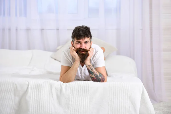 脸上开朗的男人躺在床边的白布上.卧室和床垫的概念。有胡子和胡子的马可放松一下，休息一下。躺在床上的男人，背景上的白窗帘. — 图库照片