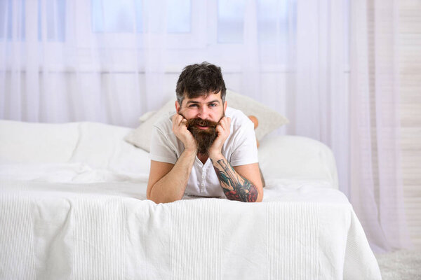 Guy on cheerful face laying on edge of bed on white sheets. Bedroom and mattress concept. Macho with beard and mustache relaxing, having rest. Man laying on bed, white curtains on background.