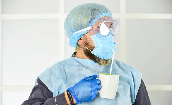 Face protection goggles mask gloves head cover. Personal protective equipment. Guy in mask drink tea coffee using straw. Infection prevention and control measures. Garments placed to protect health