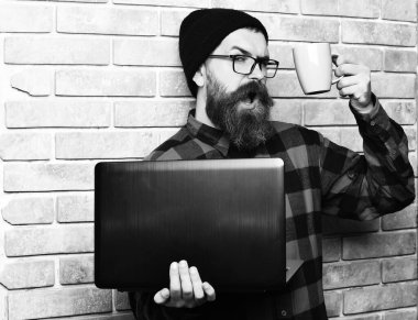 Sakallı, beyaz tenli hippi elinde bardakla dizüstü bilgisayar tutuyor.