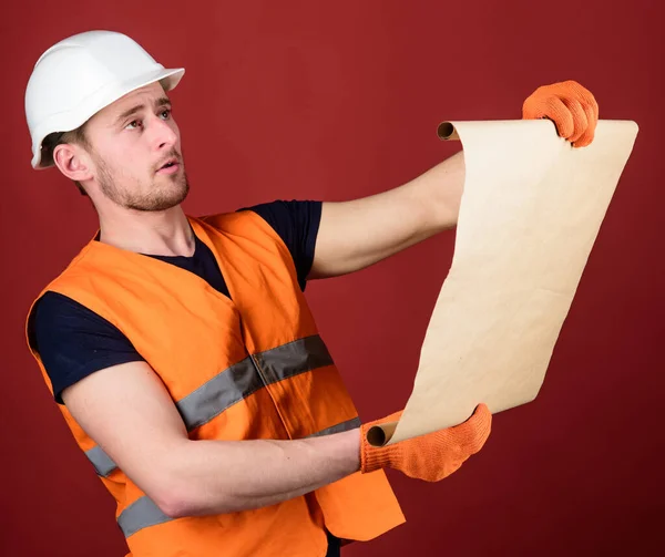 Architect concept. Engineer, architect, builder on strict face holds old blueprint in hands, supervises construction site. Man in helmet, hard hat holds building plan, controls works, red background. — Stock Photo, Image