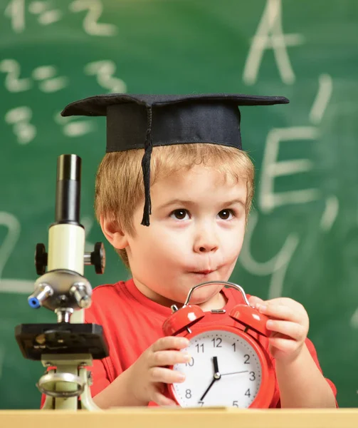 Pupil waiting for school break. School break concept. Kid boy in academic cap near microscope, holds clock in classroom, chalkboard on background. Child on excited face looks at alarm clock.