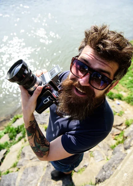 Concepto de fotógrafo reportero. Hombre con barba y bigote lleva gafas de sol, superficie de agua en el fondo. Hipster en la cara sonriente sostiene la cámara antigua. Chico disparando naturaleza cerca de río o estanque . —  Fotos de Stock