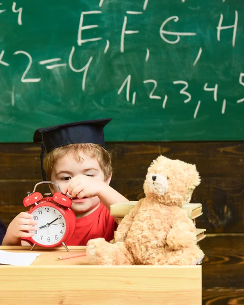 Lindo chico con gorra de graduación jugando con el reloj. Un niño aprendiendo a decir la hora. Niño rubio sentado en el escritorio en el jardín de infantes . — Foto de Stock