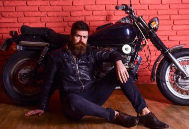 Sakallı bir adam, deri ceketli bir motorsikletli, garajda, tuğla duvarlı arka planda. Hipster, sert yüzlü, deri ceketli vahşi motorcu motosikletin yanında oturuyor. Acımasız motorcu konsepti.